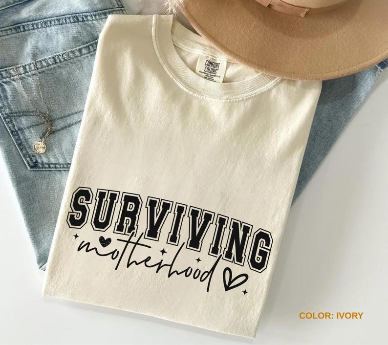 May include: Ivory-colored t-shirt with the text "SURVIVING motherhood" in black. The shirt is a crew neck and is folded on top of a pair of blue jeans. A tan hat is visible in the background.