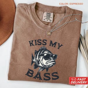 May include: Espresso-colored t-shirt with the text "KISS MY BASS" and a graphic of a fish wearing sunglasses. The shirt has a crew neck and a small tag on the collar. A silver necklace with a pendant is draped across the shirt.