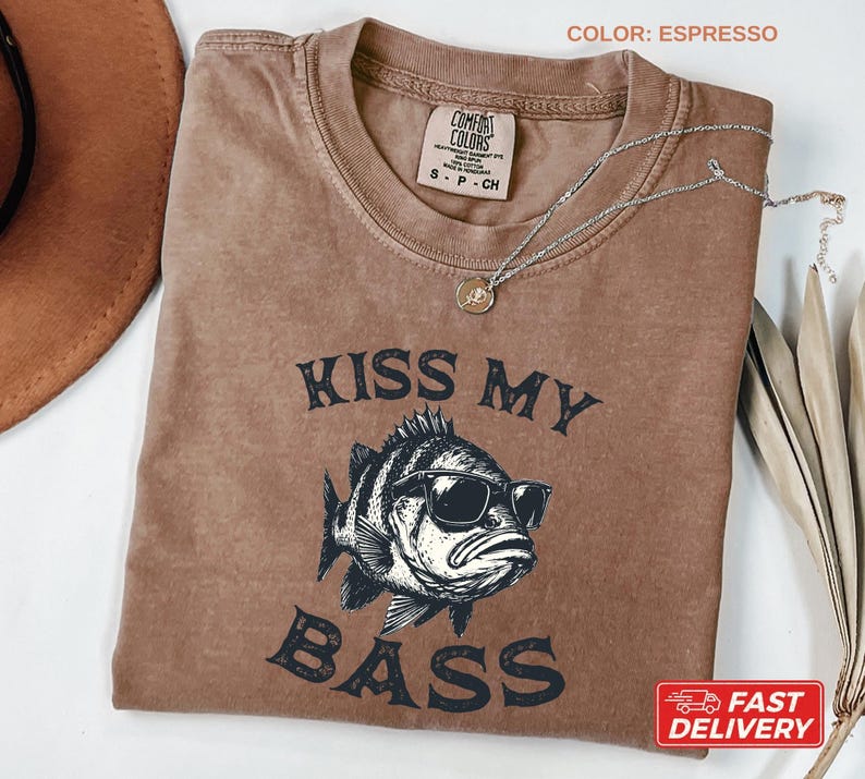 May include: Espresso-colored t-shirt with the text "KISS MY BASS" and a graphic of a fish wearing sunglasses. The shirt has a crew neck and a small tag on the collar. A silver necklace with a pendant is draped across the shirt.