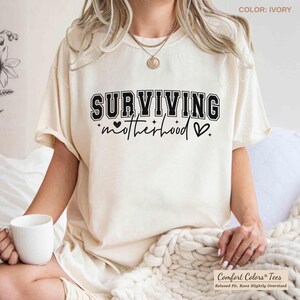 May include: Ivory-colored t-shirt with the text "SURVIVING motherhood" in black. The shirt has a relaxed fit and is slightly oversized. The text includes a heart symbol. The shirt is made by Comfort Colors.
