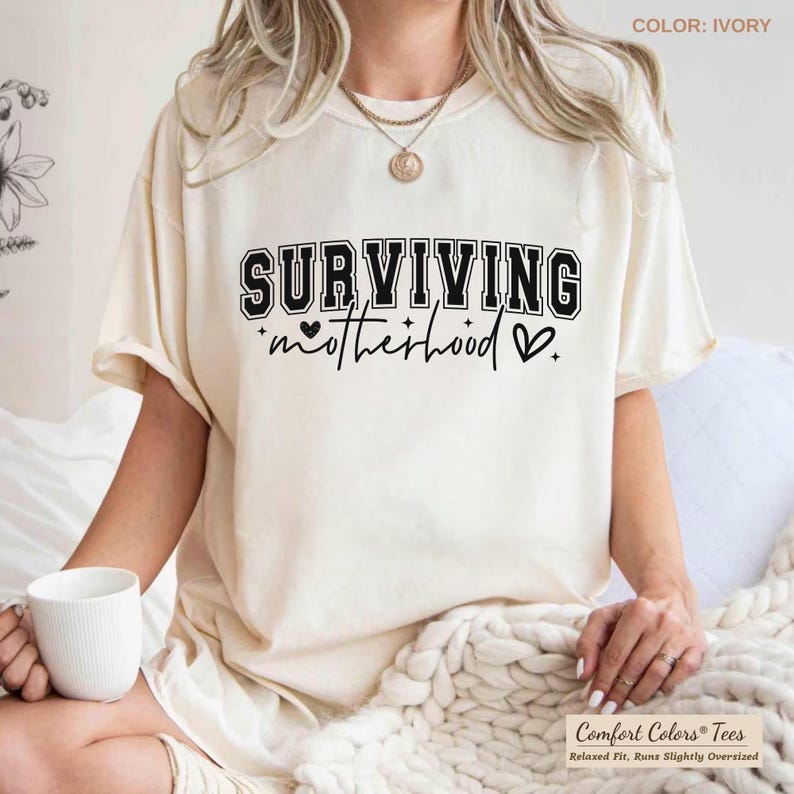 May include: Ivory-colored t-shirt with the text "SURVIVING motherhood" in black. The shirt has a relaxed fit and is slightly oversized. The text includes a heart symbol. The shirt is made by Comfort Colors.