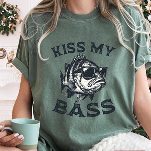 May include: Moss green t-shirt with the text "KISS MY BASS" and a graphic of a fish wearing sunglasses. The shirt has short sleeves and a relaxed fit. The shirt is made of a soft material.