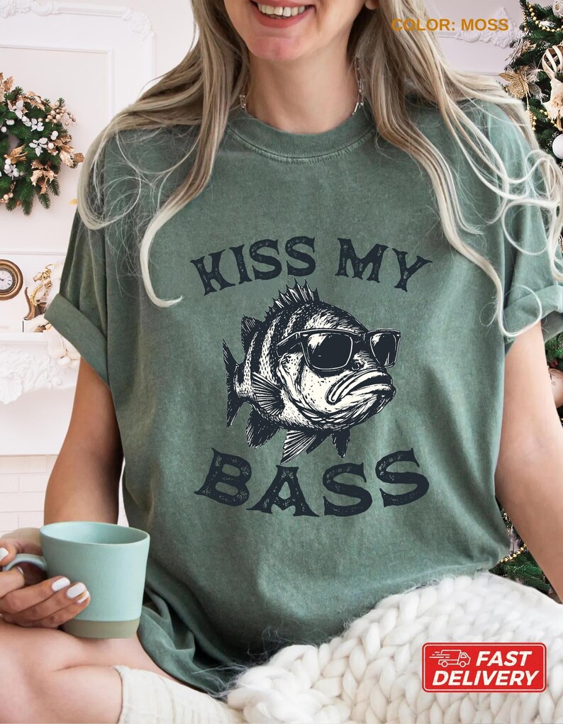 May include: Moss green t-shirt with the text "KISS MY BASS" and a graphic of a fish wearing sunglasses. The shirt has short sleeves and a relaxed fit. The shirt is made of a soft material.