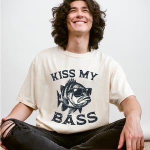 May include: Ivory-colored t-shirt featuring a graphic of a fish wearing sunglasses with the text "KISS MY BASS" in a distressed font. The shirt is a relaxed fit and has short sleeves. The image also includes a "FAST DELIVERY" sticker.