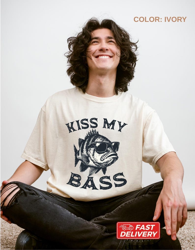 May include: Ivory-colored t-shirt featuring a graphic of a fish wearing sunglasses with the text "KISS MY BASS" in a distressed font. The shirt is a relaxed fit and has short sleeves. The image also includes a "FAST DELIVERY" sticker.