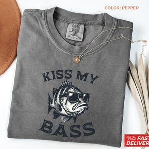 May include: Pepper-colored t-shirt with the text "KISS MY BASS" and a graphic of a fish wearing sunglasses. The shirt has a crew neck and short sleeves. A gold necklace is draped across the shirt. The shirt is made of a soft, comfortable material.