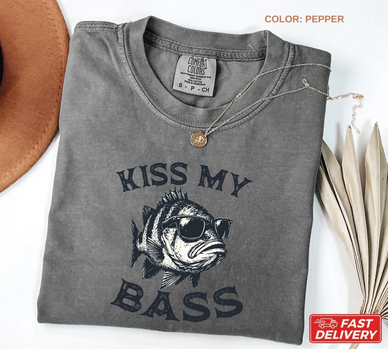 May include: Pepper-colored t-shirt with the text "KISS MY BASS" and a graphic of a fish wearing sunglasses. The shirt has a crew neck and short sleeves. A gold necklace is draped across the shirt. The shirt is made of a soft, comfortable material.