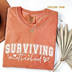 May include: A yam-colored Comfort Colors t-shirt with the words "SURVIVING motherhood" in white lettering. A gold necklace with a pendant rests on the shirt. A brown hat and dried plant are in the background.