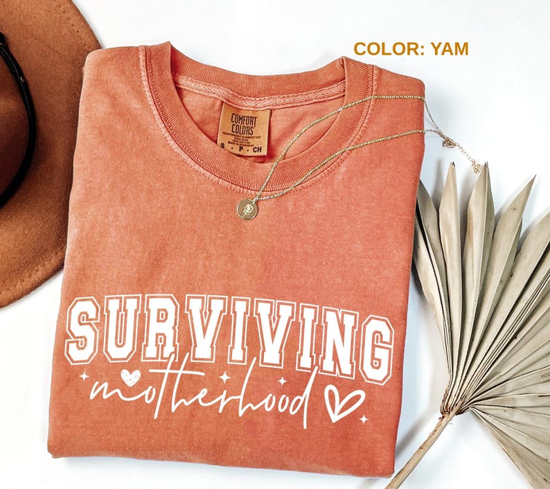 May include: A yam-colored Comfort Colors t-shirt with the words "SURVIVING motherhood" in white lettering. A gold necklace with a pendant rests on the shirt. A brown hat and dried plant are in the background.