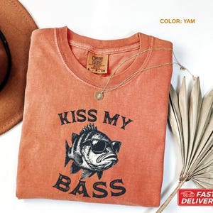 May include: A yam-colored t-shirt with the phrase "KISS MY BASS" and a graphic of a fish wearing sunglasses. The shirt has a crew neck and a small tag on the collar. A gold necklace and a brown hat are also visible.