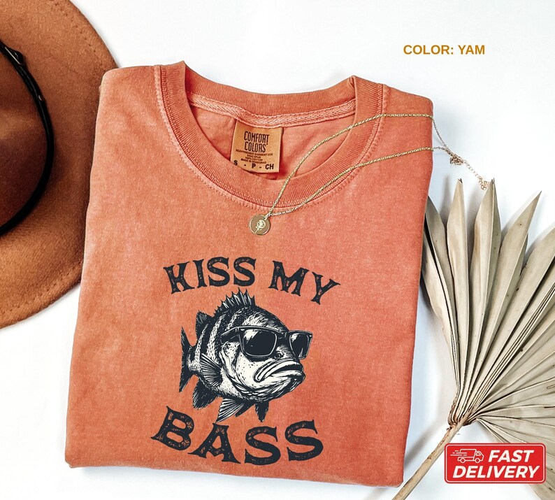 May include: A yam-colored t-shirt with the phrase "KISS MY BASS" and a graphic of a fish wearing sunglasses. The shirt has a crew neck and a small tag on the collar. A gold necklace and a brown hat are also visible.