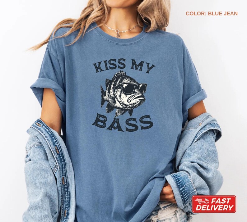 May include: Blue jean-colored t-shirt with the text "KISS MY BASS" and a graphic of a fish wearing sunglasses. The shirt has short sleeves and a relaxed fit. The color is blue jean.