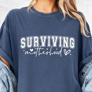 May include: Denim blue t-shirt with the text "SURVIVING motherhood" in white. The word "SURVIVING" is in a bold, collegiate-style font. The word "motherhood" is in a cursive font, with a heart and star graphic.