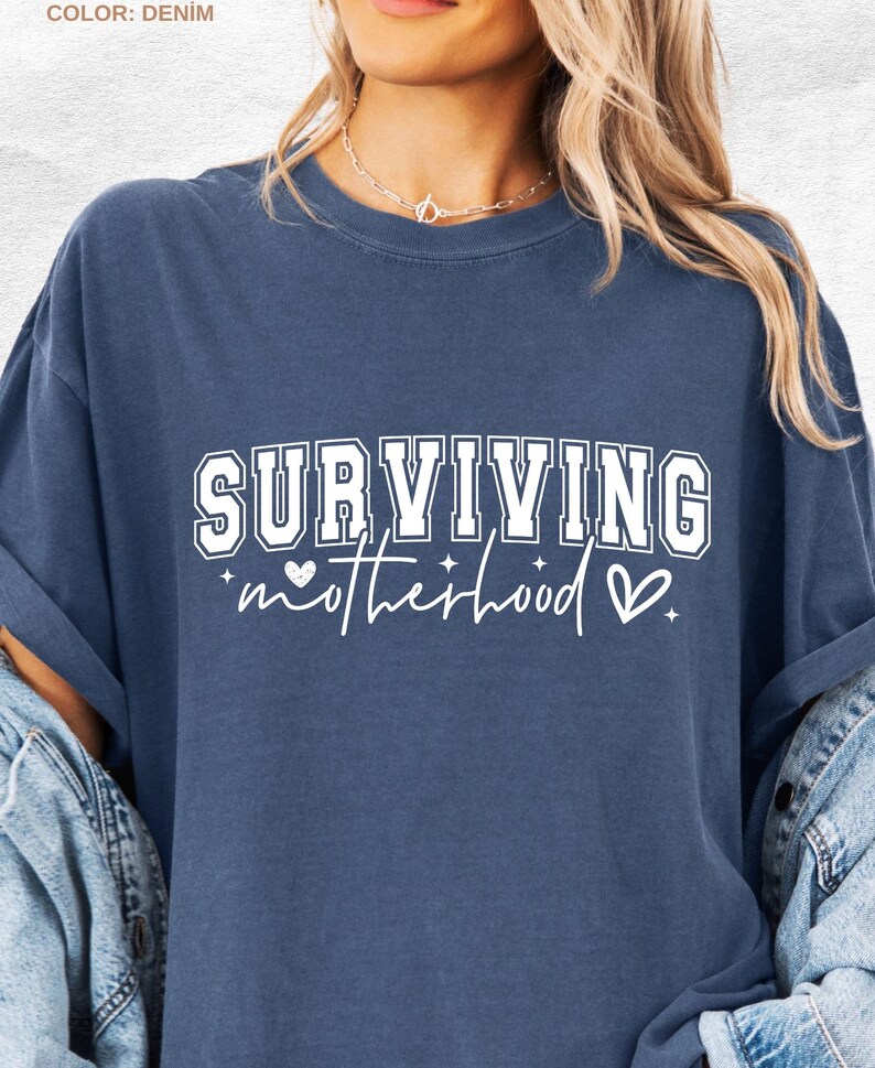 May include: Denim blue t-shirt with the text "SURVIVING motherhood" in white. The word "SURVIVING" is in a bold, collegiate-style font. The word "motherhood" is in a cursive font, with a heart and star graphic.