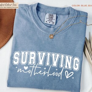 May include: Blue jean colored t-shirt with the words "SURVIVING motherhood" in white. The shirt has a crew neck and short sleeves. A gold necklace with a pendant is draped across the shirt. The shirt is folded.