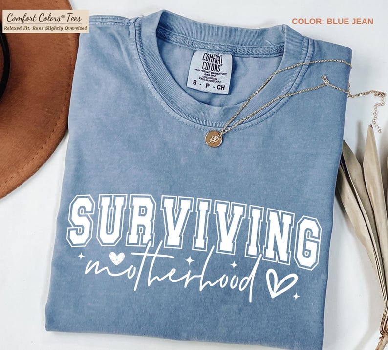 May include: Blue jean colored t-shirt with the words "SURVIVING motherhood" in white. The shirt has a crew neck and short sleeves. A gold necklace with a pendant is draped across the shirt. The shirt is folded.