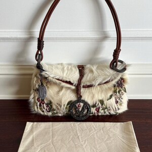 May include: Cream-colored handbag with a fur-like texture, adorned with floral embroidery in shades of red and green. Features a brown leather handle and a decorative metal charm. Includes a matching dust bag.