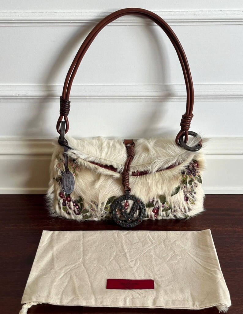 May include: Cream-colored handbag with a fur-like texture, adorned with floral embroidery in shades of red and green. Features a brown leather handle and a decorative metal charm. Includes a matching dust bag.