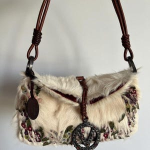 May include: Cream-colored fur handbag with brown leather straps and accents. The bag features floral embroidery in shades of pink, green, and purple. A metal charm with the letter "V" hangs from the front. The bag has a rectangular shape.