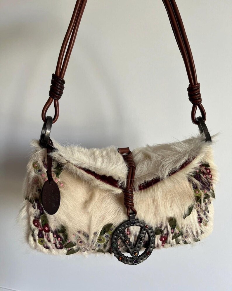 May include: Cream-colored fur handbag with brown leather straps and accents. The bag features floral embroidery in shades of pink, green, and purple. A metal charm with the letter "V" hangs from the front. The bag has a rectangular shape.