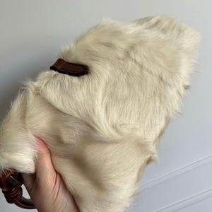 May include: Cream-colored, furry handbag with a brown leather accent and handle. The bag has a soft, textured appearance. The handle is a dark brown, possibly wooden, ring.