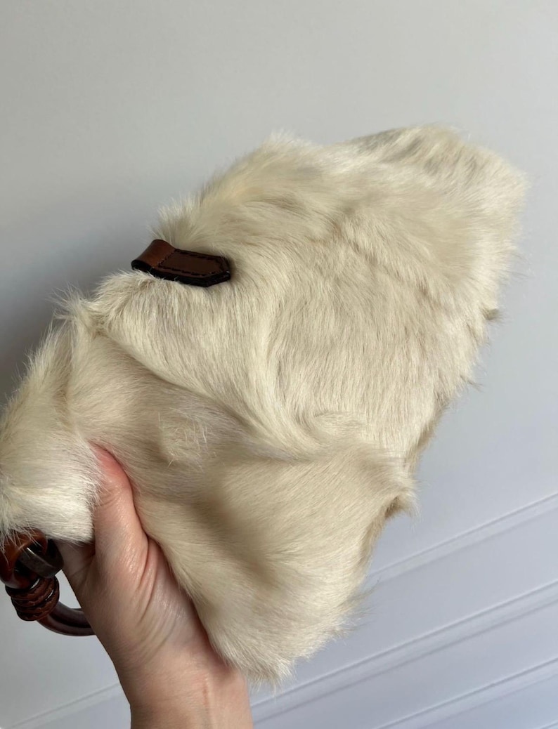 May include: Cream-colored, furry handbag with a brown leather accent and handle. The bag has a soft, textured appearance. The handle is a dark brown, possibly wooden, ring.