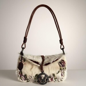 May include: Cream-colored fur handbag with brown leather straps. The bag features floral embroidery in shades of purple, green, and red. A metal charm with the letter "V" hangs from the front. The bag has a flap closure.