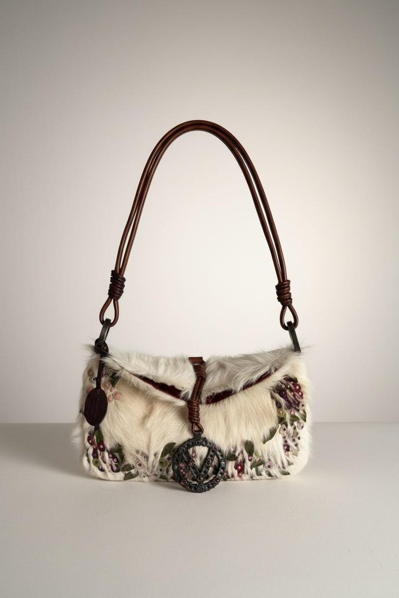 May include: Cream-colored fur handbag with brown leather straps. The bag features floral embroidery in shades of purple, green, and red. A metal charm with the letter "V" hangs from the front. The bag has a flap closure.