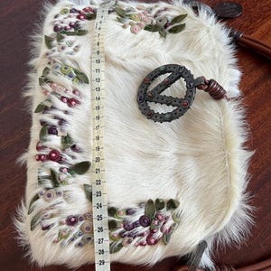 May include: Cream-colored, furry handbag with floral embroidery in pink, green, and purple. The bag features a decorative metal clasp and a brown leather strap. The bag measures approximately 33 inches in length.