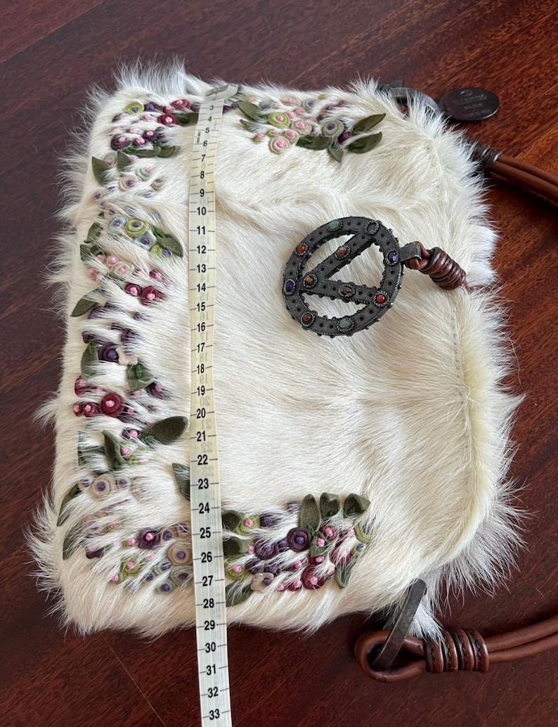 May include: Cream-colored, furry handbag with floral embroidery in pink, green, and purple. The bag features a decorative metal clasp and a brown leather strap. The bag measures approximately 33 inches in length.