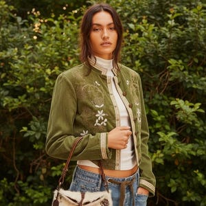 May include: Olive green jacket with floral embroidery and gold trim. The jacket is paired with a white turtleneck, blue jeans, and a small handbag with a brown leather strap and a patterned fur design.