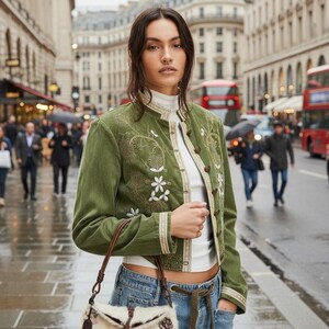 May include: A woman in a green jacket with floral embroidery and a small, patterned handbag stands on a city street. The jacket has a mandarin collar and button closures. She wears a white turtleneck and blue jeans. The background shows buildings and red double-decker buses.