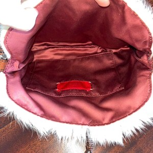 May include: Open view of a small, burgundy-colored handbag with a white, furry trim. The interior features a burgundy satin lining and a small pocket with a red tag. A decorative silver pendant hangs from the bag.