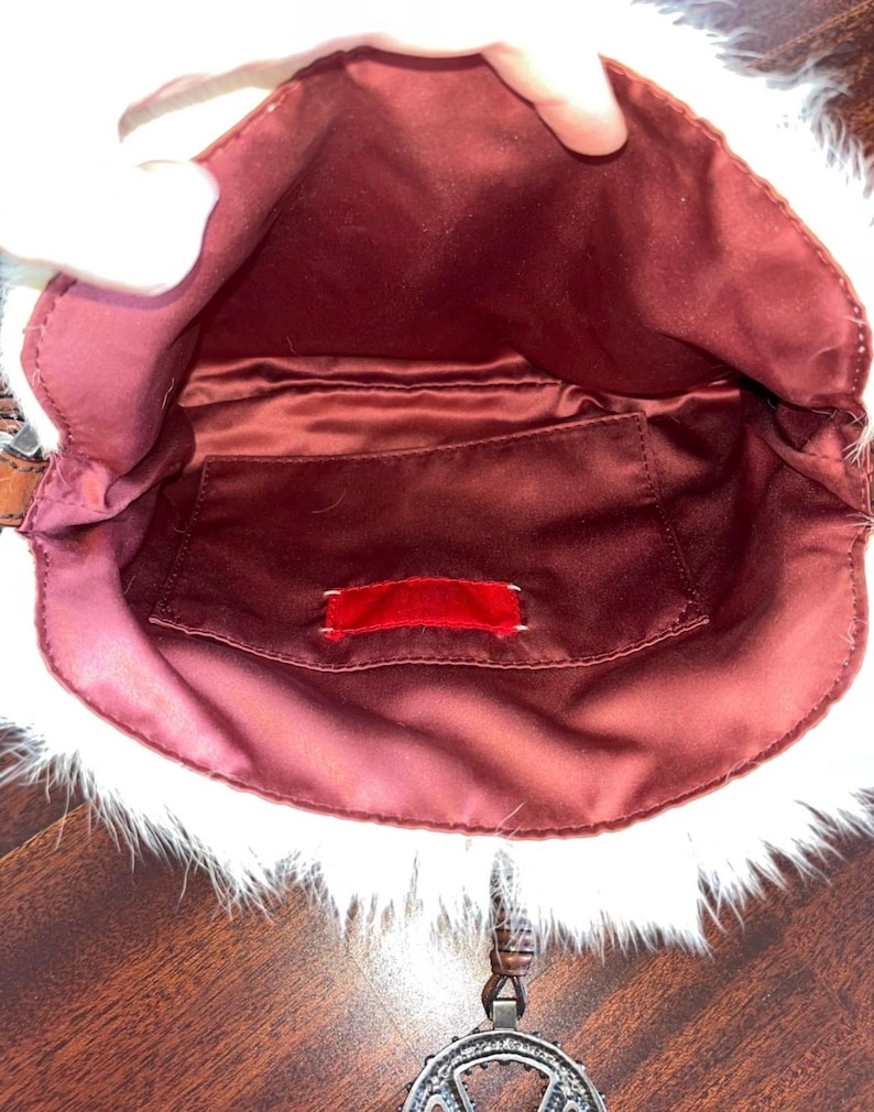 May include: Open view of a small, burgundy-colored handbag with a white, furry trim. The interior features a burgundy satin lining and a small pocket with a red tag. A decorative silver pendant hangs from the bag.