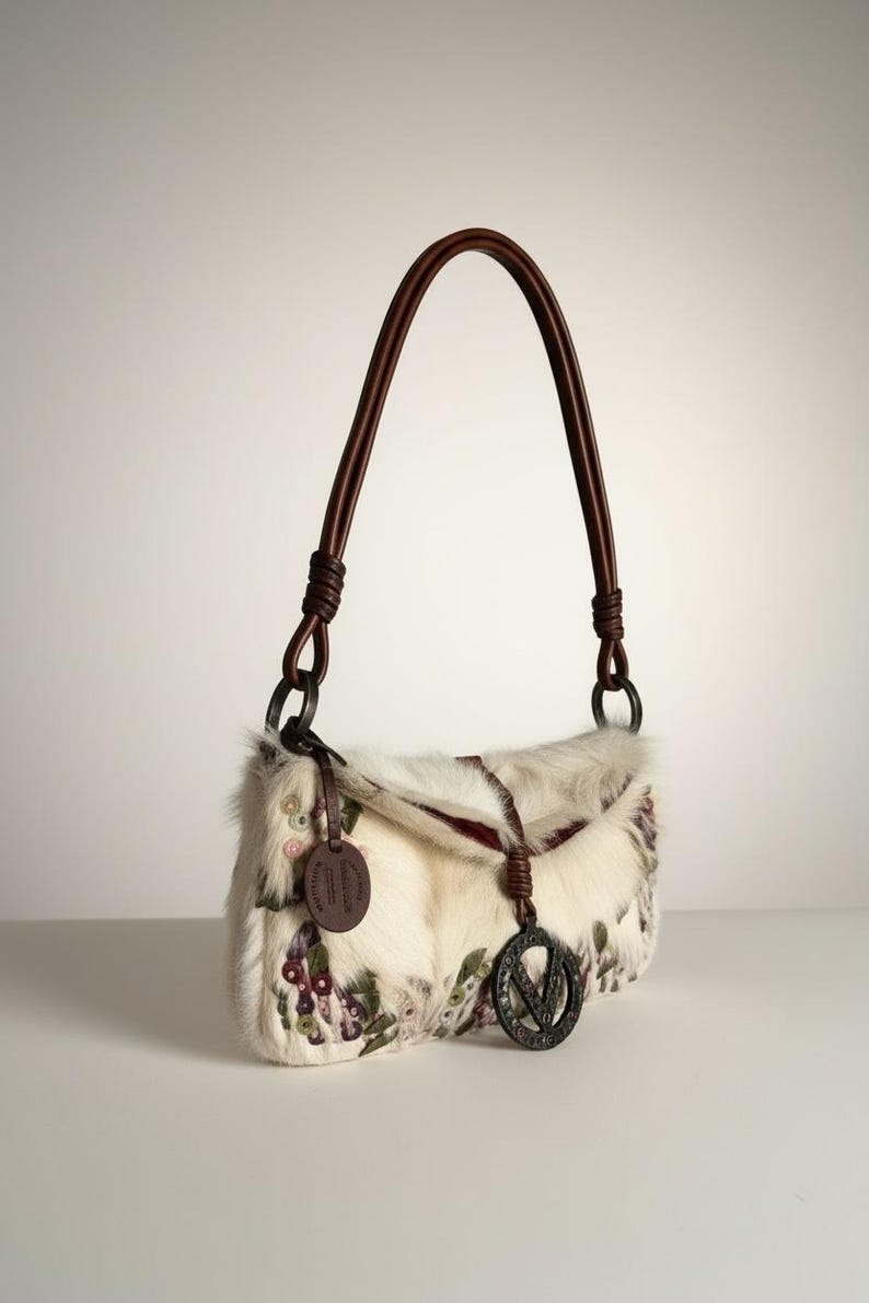 May include: Cream-colored handbag with a brown leather handle. The bag features a floral pattern with purple and green accents. A brown leather tag and a metal charm with a "V" are attached to the bag.