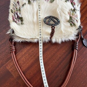 May include: Cream-colored handbag with a fur-like texture, adorned with floral embellishments in pink, green, and purple. The bag features a metal clasp and brown leather handles. The bag measures approximately 16 inches in length.