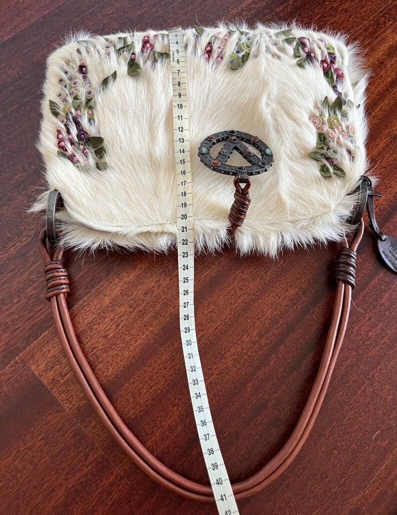 May include: Cream-colored handbag with a fur-like texture, adorned with floral embellishments in pink, green, and purple. The bag features a metal clasp and brown leather handles. The bag measures approximately 16 inches in length.