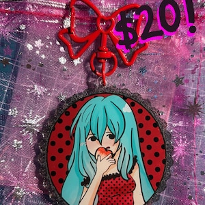 May include: A red bow-shaped keyring with a circular pendant featuring an anime-style illustration of a person with blue hair eating an apple. The pendant has a red background with black polka dots and a black lace trim.