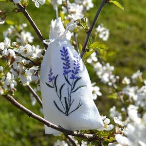 Può includere: Un sacchetto di lino bianco con ricamo di lavanda. Il sacchetto è appeso a un ramo di un albero fiorito con fiori bianchi.