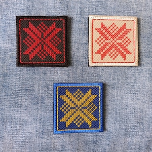 Op de afbeelding: Drie vierkante geborduurde patches met een geometrisch kruissteekpatroon. Eén patch is zwart met rode stiksels, één is beige met rode stiksels en één is blauw met gele stiksels. Elke patch is voorzien van een bijpassende gestikte rand.