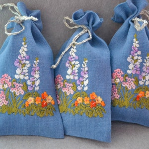 Pode incluir: Três pequenas bolsas de linho azul com desenhos florais bordados. As flores são em tons de rosa, roxo, branco, laranja e verde.