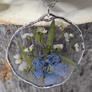 Puede incluir: Collar plateado con un colgante que contiene flores secas encapsuladas en resina transparente. El colgante tiene un borde plateado irregular. Las flores incluyen una gran flor azul y pequeñas flores blancas.