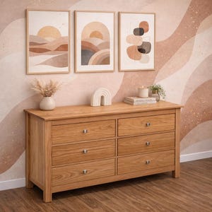 Könnte beinhalten: Eine hellbraune Holzkommode mit sechs Schubladen und silberfarbenen Knöpfen. Darüber befinden sich drei gerahmte abstrakte Kunstdrucke in neutralen Tönen, ein weißer Keramikregenbogen und dekorative Gegenstände. Die Wand hat ein wellenförmiges Muster.