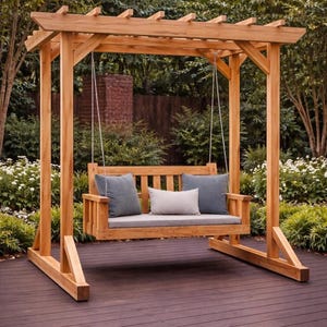 Op de afbeelding: Een houten schommel met een pergola-achtig dak. De schommel heeft een houten zitting met grijze kussens en kussens. De constructie is gemaakt van lichtgekleurd hout en staat op een donkerbruin terras, omringd door groen.