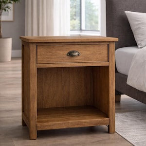 May include: A wooden nightstand with a drawer and open shelf. The nightstand is made of light brown wood and has a dark metal handle on the drawer. The nightstand is next to a bed.