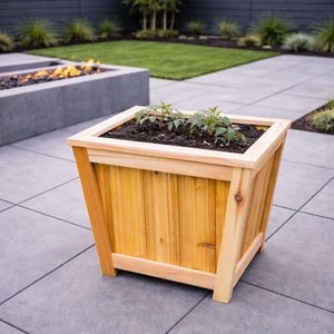 May include: A wooden planter box with a light brown finish, filled with soil and small green plants. The box has a square shape with angled sides and is set on a gray tiled surface, suggesting outdoor use for gardening.