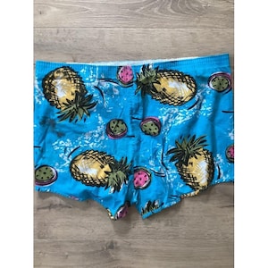 Könnte beinhalten: Hellblaue Badehose mit einem sich wiederholenden Muster aus gelben Ananas mit grünen Blättern und aufgeschnittenen rosa Drachenfrüchten. Der Stoff weist ein weißes Spritzmuster auf dem gesamten Design auf, mit einem elastischen Bund.
