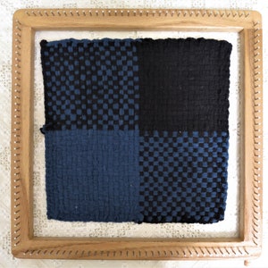 May include: A wooden loom with a woven fabric in shades of blue and black. The fabric is divided into four squares, each with a different pattern.