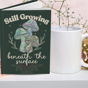 May include: A dark green hardcover journal featuring a graphic of three textured mushrooms in shades of purple, blue, and green. The text reads Still Growing beneath the surface in a cream font, accompanied by white line art of roots and leaves.