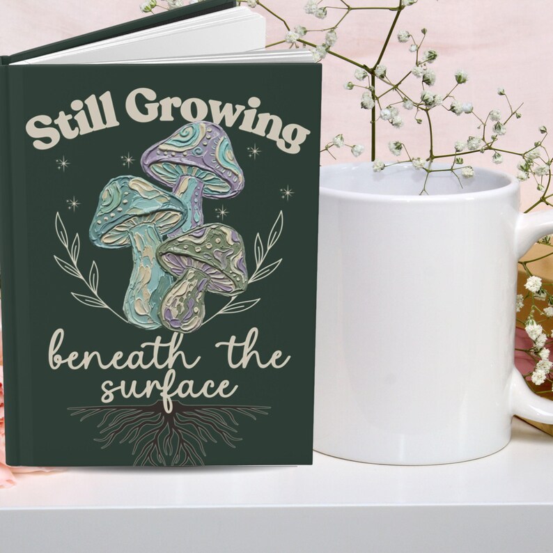 May include: A dark green hardcover journal featuring a graphic of three textured mushrooms in shades of purple, blue, and green. The text reads Still Growing beneath the surface in a cream font, accompanied by white line art of roots and leaves.